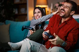 Couple laughing while playing video games together on a sofa in their living room.